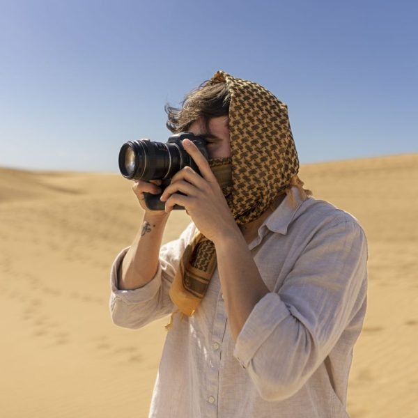 Person wearing a patterned head scarf and light shirt, taking a photograph with a DSLR in a sandy desert landscape.